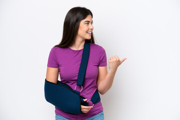 Young Brazilian woman with broken arm and wearing a sling isolated on white background pointing to...