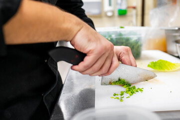 A close-up of a chef's hands skillfully chopping lettuce with a sharp knife on a cutting board in a professional kitchen setting. Fresh ingredients are visible in the background.