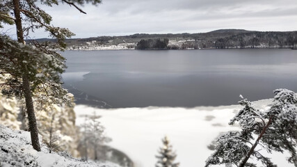 a winter landscape where a beautiful lake stretches across the scene. The center of the lake is still open water, reflecting the gray winter sky and the snow-covered trees that line its edges. On one 