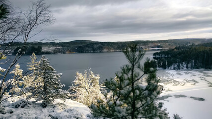 a winter landscape where a beautiful lake stretches across the scene. The center of the lake is still open water, reflecting the gray winter sky and the snow-covered trees that line its edges. On one 