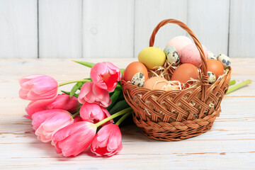 Eggs in a wicker basket and bouquet of pink tulips on white wooden table.