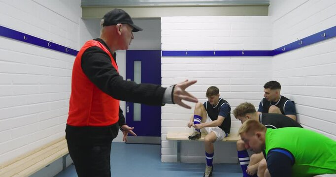 Male soccer players wearing black vests and tying shoes sitting on bench in changing room with coach