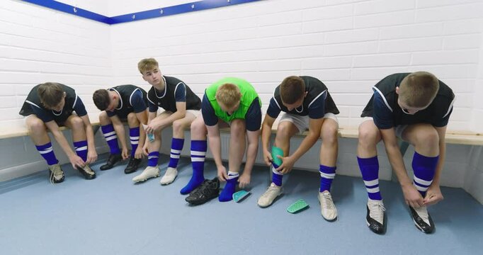 Male soccer players wearing black vests and tying shoes sitting on bench in changing room