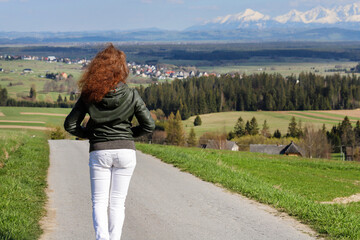 A young redhead girl walks along a scenic road in the mountains. Around her are beautiful meadows and forests.