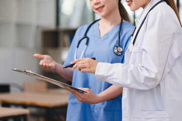 Two healthcare professionals, one in white coat and other in blue scrubs, are discussing patient information while reviewing clipboard in bright medical setting