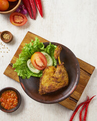 indonesian traditional ayam greprek, a fried chicken with sambal chili sauce on top of it, on a white wooden background.