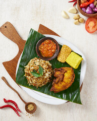 Indonesian fried chicken with spiced rice, tempe and tofu, on top of banana leaves, in a wooden background