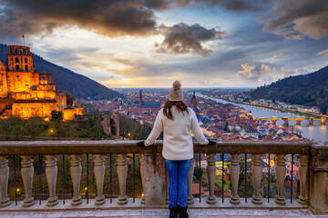 A tourist woman enjoys the winter sunset view of the illuminated city skyline of Heidelberg, Germany, with castle and river Neckar