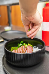 A hand adds fresh herbs to a bowl of noodles topped with sliced beef. The dish is presented in a stylish black bowl, showcasing vibrant colors and textures in a kitchen setting.