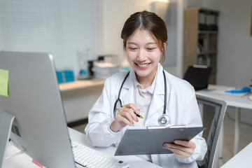 Young female physician smiling and working with computer and taking notes on clipboard in her bright modern office