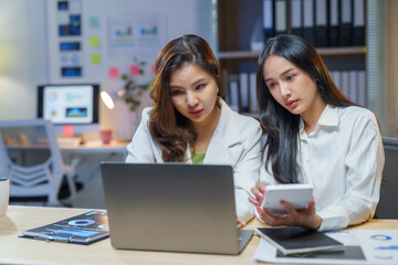 Two businesswomen analyzing financial data on a laptop and using a calculator, working late in the office