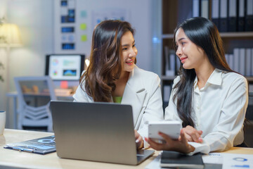 Two Asian businesswomen working together using laptop and calculator in office at night