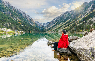 Fototapeta premium A serene moment at Lac de Gaube, French Pyrenees, where crystal-clear waters reflect towering peaks, vibrant greenery, and a traveler in a striking red coat.