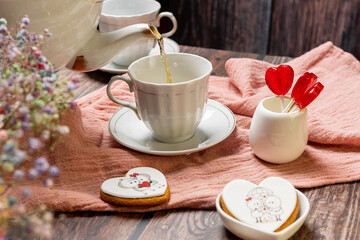 a woman pours tea from a white teapot into a white porcelain cup with a saucer, next to gingerbread cookies with white glaze, red heart-shaped lollipops, Valentine's Day composition