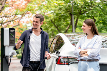 Lovely young couple recharging battery for electric car during autumnal road trip travel EV car in autumnal forest. Eco friendly travel on vacation during autumn. Exalt