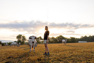 Obraz premium Young female farmer standing in the pasture with her grazing cows at sunrise