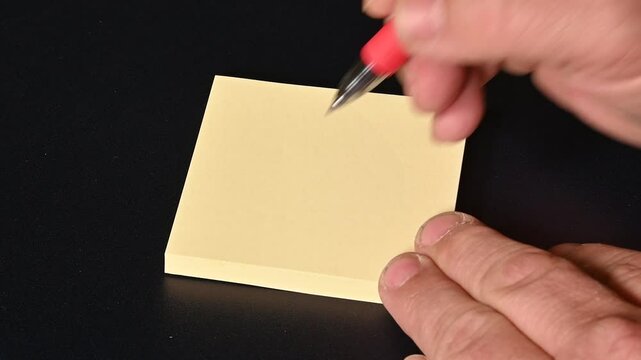 A hand holding a blue pen writes on a yellow sticky note (post-it), creating two dots and a curved upward line that form a smiling face (smiley). The composition is square-shaped and set against 