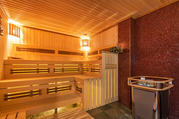 Modern sauna design made of light wood. Benches and headrest, stylish design of the heating element against the wall decorated with mosaic tiles.
