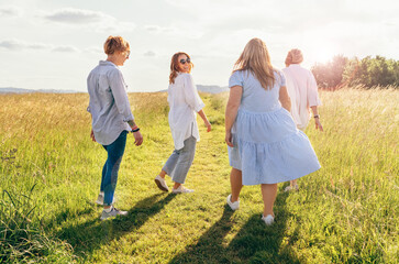 Naklejka premium Portrait of four cheerful smiling women holding hand in hand walking by a high green grass meadow. They looking at the camera. Woman's friendship, relations, and happiness concept image.