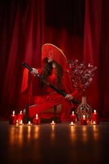 Beautiful young girl in a red kimono dress with a katana on red curtains background..