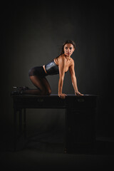 Stunning girl in black tights stockings and leather black corset posing on an old table on gray...