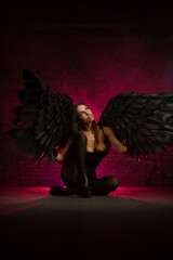 Beautiful girl in a black corset and black tights with black angel wings on pink smoky background.
