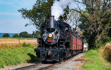 A classic locomotive puffs white smoke as it moves along rural tracks surrounded by golden cornfields and green trees on a sunny autumn afternoon.
