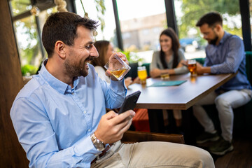 Businessman using his phone in the restaurant. Smiling and drinking whisky.