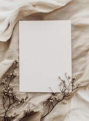Blank white invitation rests on top of soft beige fabric, with delicate flowers and leaves along the bottom edge. The overall aesthetic is minimalist, with a neutral color palette