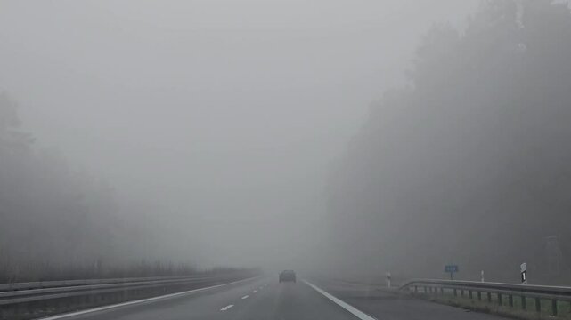 Ein Auto f&auml;hrt auf einer nebligen Autobahn in Deutschland. Die dichte Nebeldecke verdeckt die Sicht, w&auml;hrend kahle B&auml;ume die Stra&szlig;e s&auml;umen