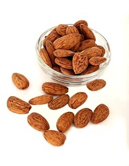 The photo shows a glass bowl of almonds in their shells, placed on a white background. Some of the nuts are scattered nearby, creating a natural composition.