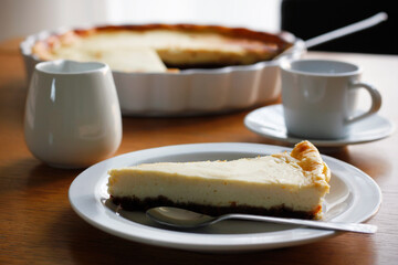 Piece of homemade cheesecake on a white plate on wooden table, a cup of black coffee with saucer, a cup of milk and a baking dish in the background. Coffee nad cake, side view, selective focus.