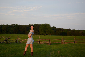Fototapeta premium Sexy woman in short and tight blue sundress and boots stands in fenced in pasture - rural or farm