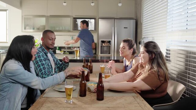 Diverse group of happy male and transgender female friends drinking beer, cheering in living room. friends hanging out at home enjoying leisure time together.
