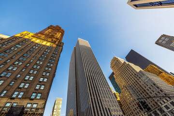 Towering skyscrapers in Midtown Manhattan reflecting golden sunlight against clear blue sky on bright spring afternoon.