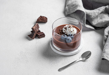 Chocolate pudding in a glass with whipped cream and fresh blueberry on a light background