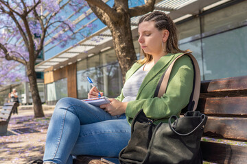 Young woman writing in a notebook while sitting on a bench under blooming jacaranda trees.