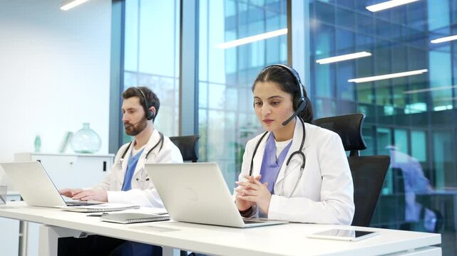 A team of two doctors in headset working in online call center healthcare service using laptop. Medical workers professionals man and woman talking consulting patient by video call sitting in office