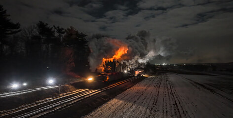A train car is on fire next to snowy fields under a moonlit sky. Smoke billows into the air as...