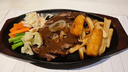 Grilled beef steak served with creamy brown gravy crispy potato wedges french fries and sautéed vegetables on a black sizzling plate creating a delicious hearty meal presentation.