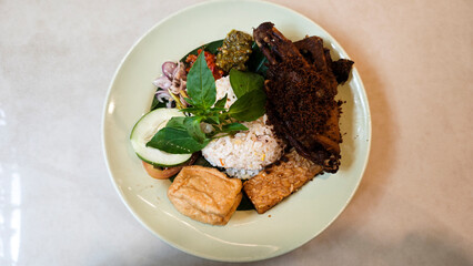 Traditional Indonesian Kecombrang Rice with Fried Spiced Duck Tempeh Tofu Assorted Sambals Fresh Basil and Cucumber Served on a Light Green Plate on Neutral Table Background