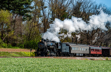 A classic steam locomotive pulls vintage passenger cars along a narrow track surrounded by vibrant green fields and trees under a clear blue sky.