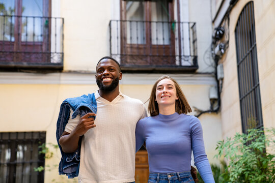 Happy multiracial couple walking out of their building to experience the lively streets. Concept of urban joy and connection. - Powered by Adobe