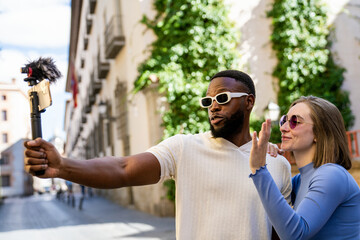 Multiethnic couple of influencers recording a video blog on a picturesque city street, using a smartphone with an external microphone