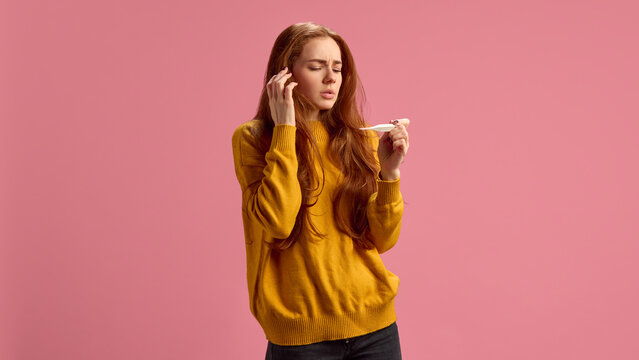 Young woman in her 20s feeling sick, measuring temperature, suffering from fever during seasonal cold on pink studio background. Concept of illness, medicine, treatment, health care