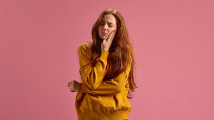 Young red-haired woman suffering from exhausting toothache, holding hand on her cheek on pink...