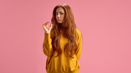 Fototapeta premium Young red-haired woman in yellow sweater holding pill on pink studio background. Taking meds, vitamins. Concept of illness, medicine, treatment, health care