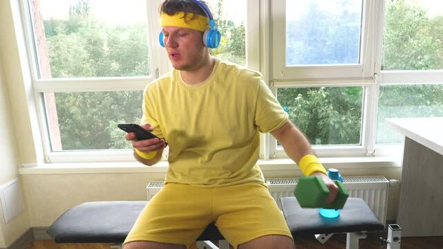 A comedian athlete with dumbbells pumps a muscle while listening to music on headphones. A young funny guy training at home. Workout with dumbbells. Slow motion.