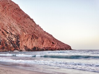 Ein roter Berg am Meer mit blauen Wellen und ein Strand mit beigem Sand
