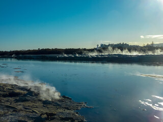 Fototapeta premium Geothermal area with steam rising from vents and hot springs along shoreline of Rotorua lake, Bay of Plenty Region, North Island New Zealand. Sky is clear blue. mineral-rich wet sand and mudflats